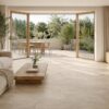 Photo d’un salon contemporain lumineux avec sol en grands carreaux de grès cérame effet travertin, baie vitrée ouverte sur une terrasse assortie, mobilier bois clair, canapé crème, plantes discrètes. Ambiance chaleureuse, décoration naturelle, lumière de fin de matinée