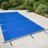 Bâche de piscine bleue tendue sur une terrasse en pierre claire, bordée d’un jardin de végétation dense et verdoyante, avec pots de plantes et sangles de fixation visibles