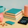 Scène colorée avec tirelire rose, calculatrice, pièces de monnaie et livres empilés sur une table jaune, évoquant le budget maison 2025-26