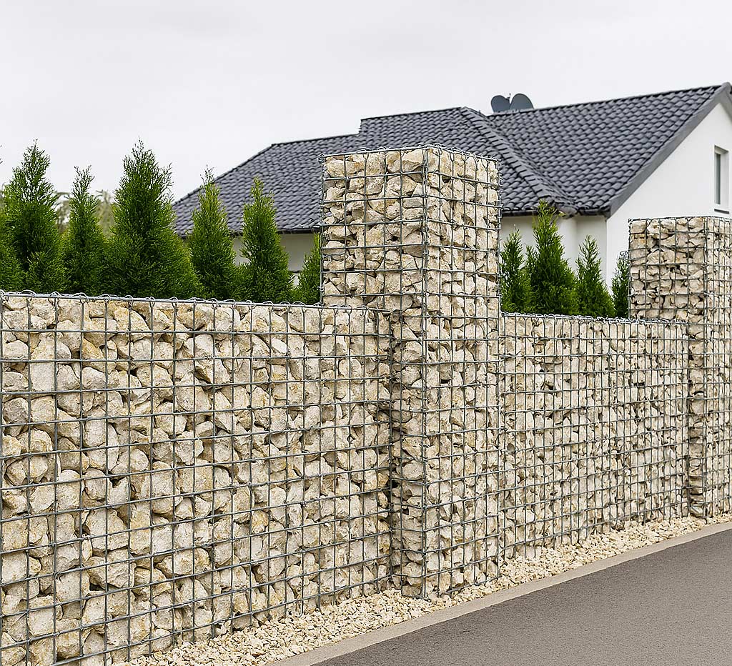 Photographie en haute résolution montrant une clôture en gabion composée de cages métalliques galvanisées remplies de pierres claires, ponctuée de piliers massifs à intervalles réguliers. Derrière la clôture, une haie dense de thuyas forme un écran végétal devant une maison moderne aux murs blancs et au toit en tuiles anthracite, le tout sous un ciel nuageux.