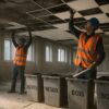 Curage manuel : 2 opérateurs en EPI (gilet HV orange, casque bleu), dépose de faux-plafond et menuiseries, bennes étiquetées “INERTES/MÉTAUX/BOIS”