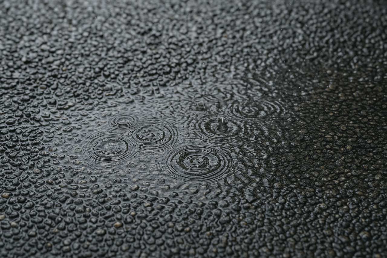 Gros plan d’une surface de béton drainant en pente avec gouttes de pluie s’infiltrant, texture poreuse bien visible, bordure pierre, ruissellement maîtrisé, lumière douce