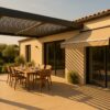 Terrasse contemporaine en été, maison claire, grande pergola bioclimatique adossée avec lames partiellement ouvertes, table dressée pour 6, store banne voisin déployé sur baie vitrée, ombre nette au sol, ambiance dorée de fin d’après-midi