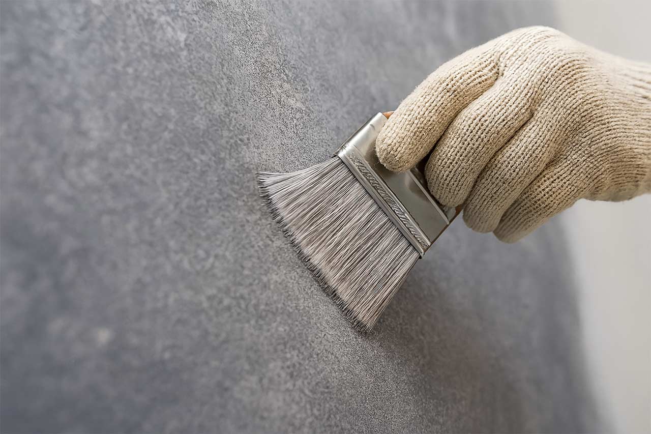 Main gantée appliquant une peinture effet béton ciré sur un mur à l’aide d’un large pinceau plat, illustrant une étape de finition décorative murale avec texture minérale grise.