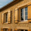 Vue en contre-plongée d’une façade de maison française en pierre, avec plusieurs volets battants en bois fraîchement rénovés, ferrures noires neuves bien visibles (gonds, espagnolettes, butées)