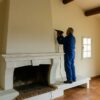 Ouvrier en combinaison bleue utilisant une perceuse pour travailler au-dessus d’une cheminée en stuc blanc dans un salon lumineux avec poutres apparentes, carrelage en terre cuite et grande baie vitrée donnant sur l’extérieur.