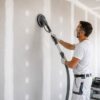Plaquiste ponçant un mur en plaques de plâtre avec une ponceuse reliée à un aspirateur.