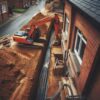 La photographie montre un chantier de raccordement au tout-à-l'égout adjacent à une maison en briques. L'angle de vue est légèrement en plongée, capturant une pelleteuse en action creusant une tranchée le long de la façade.